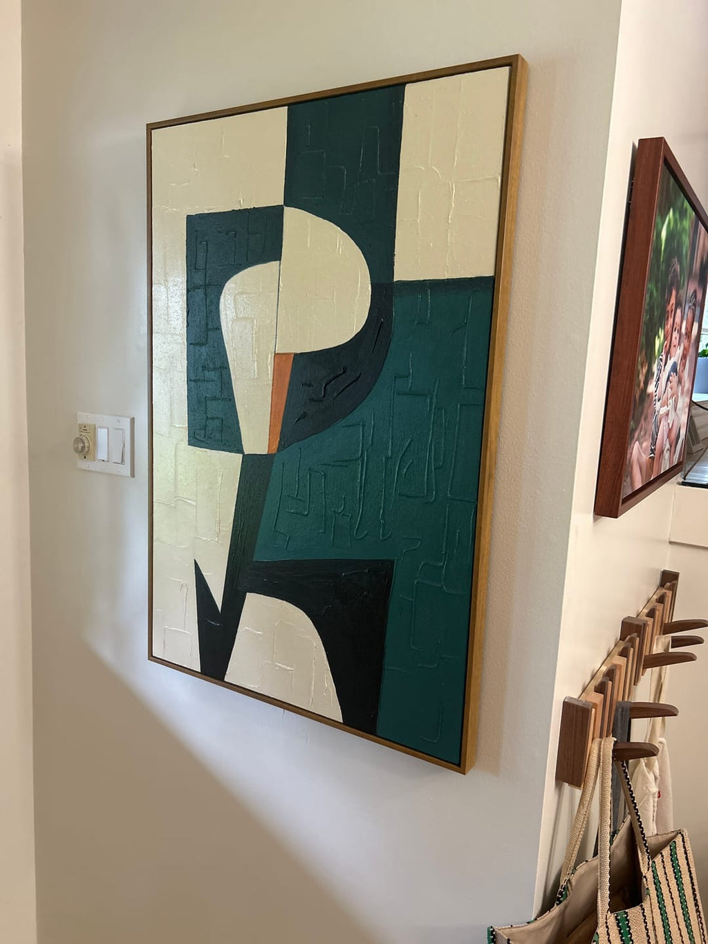 A large abstract geometric wall art painting with a textured, raised canvas surface, featuring blocks of teal, cream, black, and a small orange accent, framed in a light wood border and hung on a white wall.