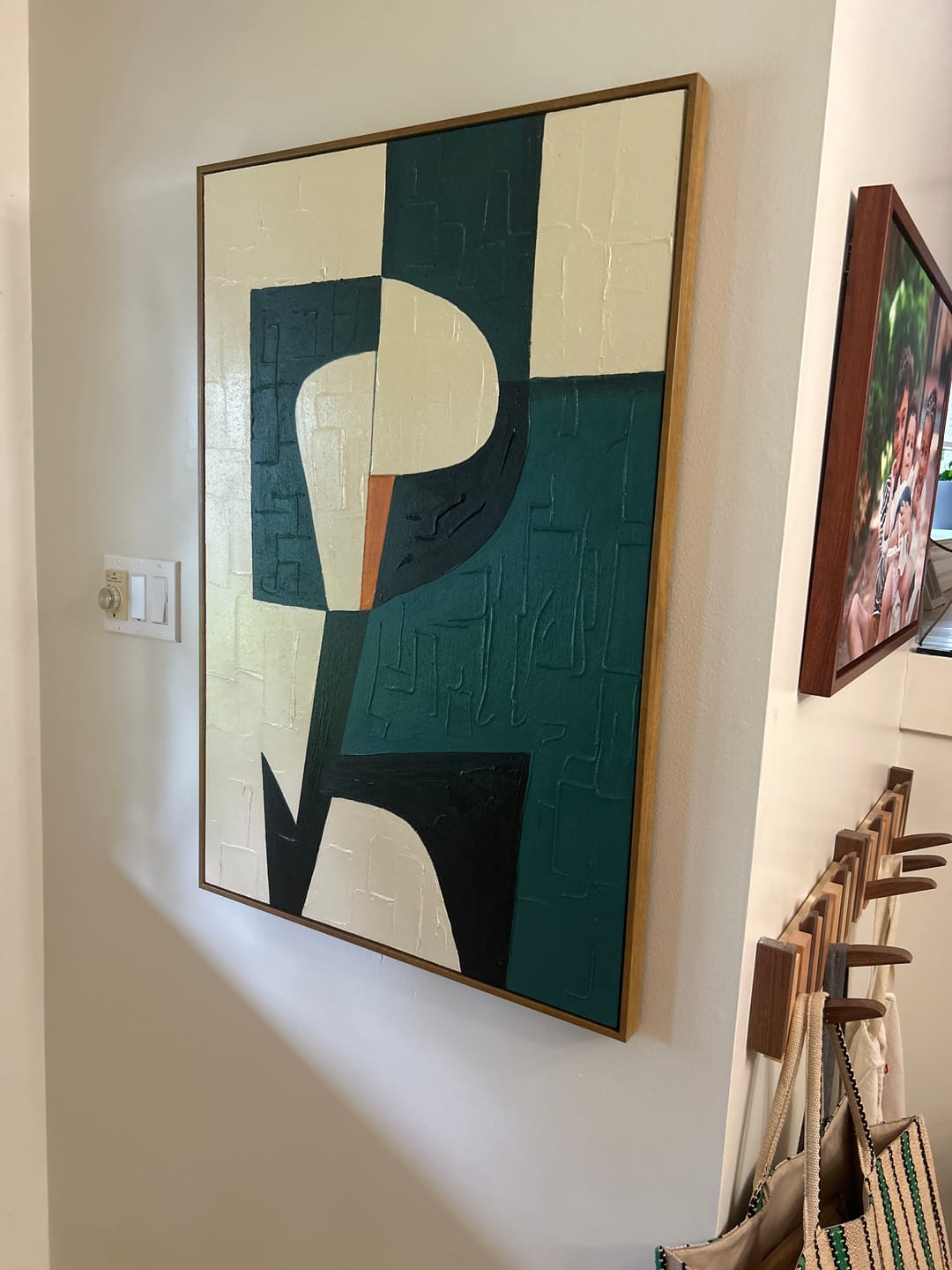 A large abstract geometric wall art painting with a textured, raised canvas surface, featuring blocks of teal, cream, black, and a small orange accent, framed in a light wood border and hung on a white wall.