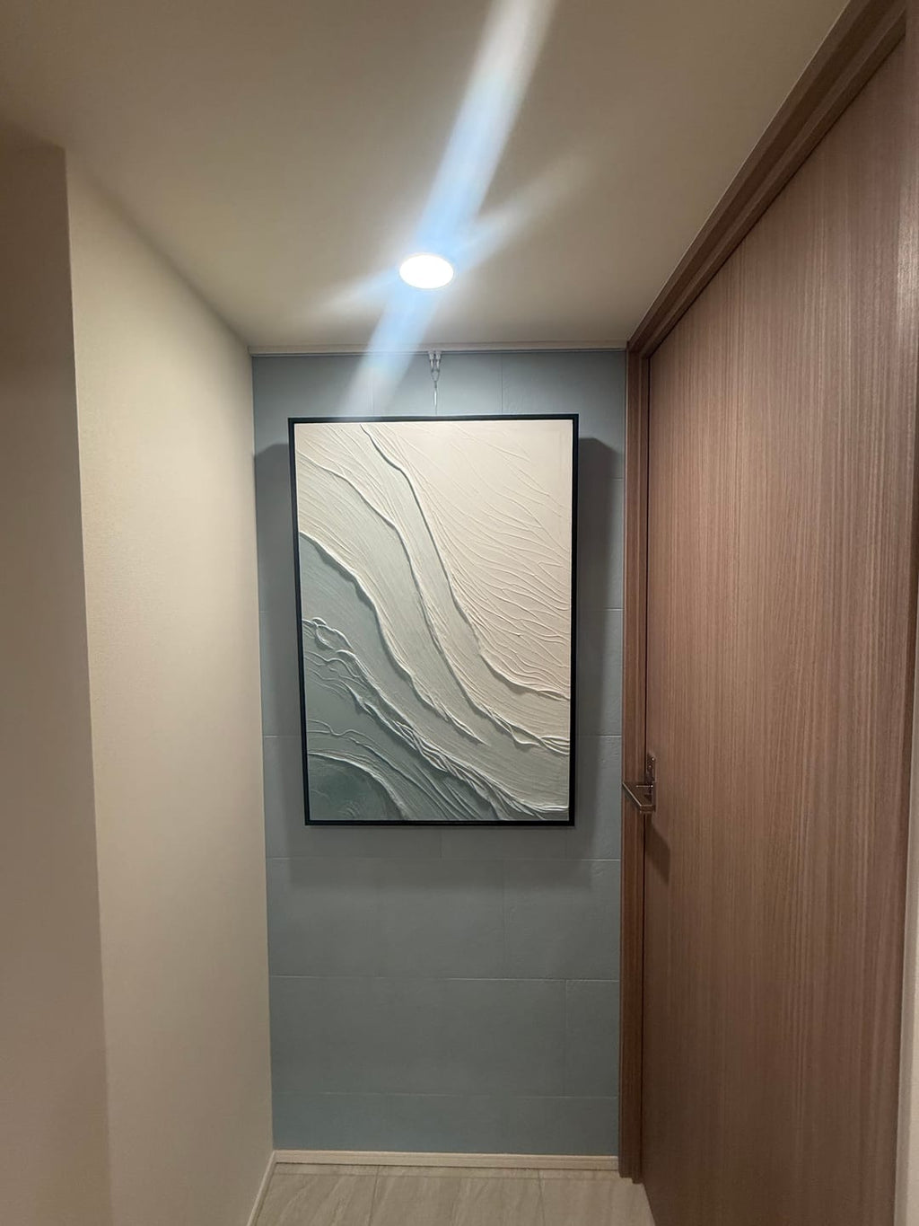 A large framed wabi sabi abstract wall art print with flowing white and gray textured lines, displayed on a light blue-gray tiled wall in a hallway.