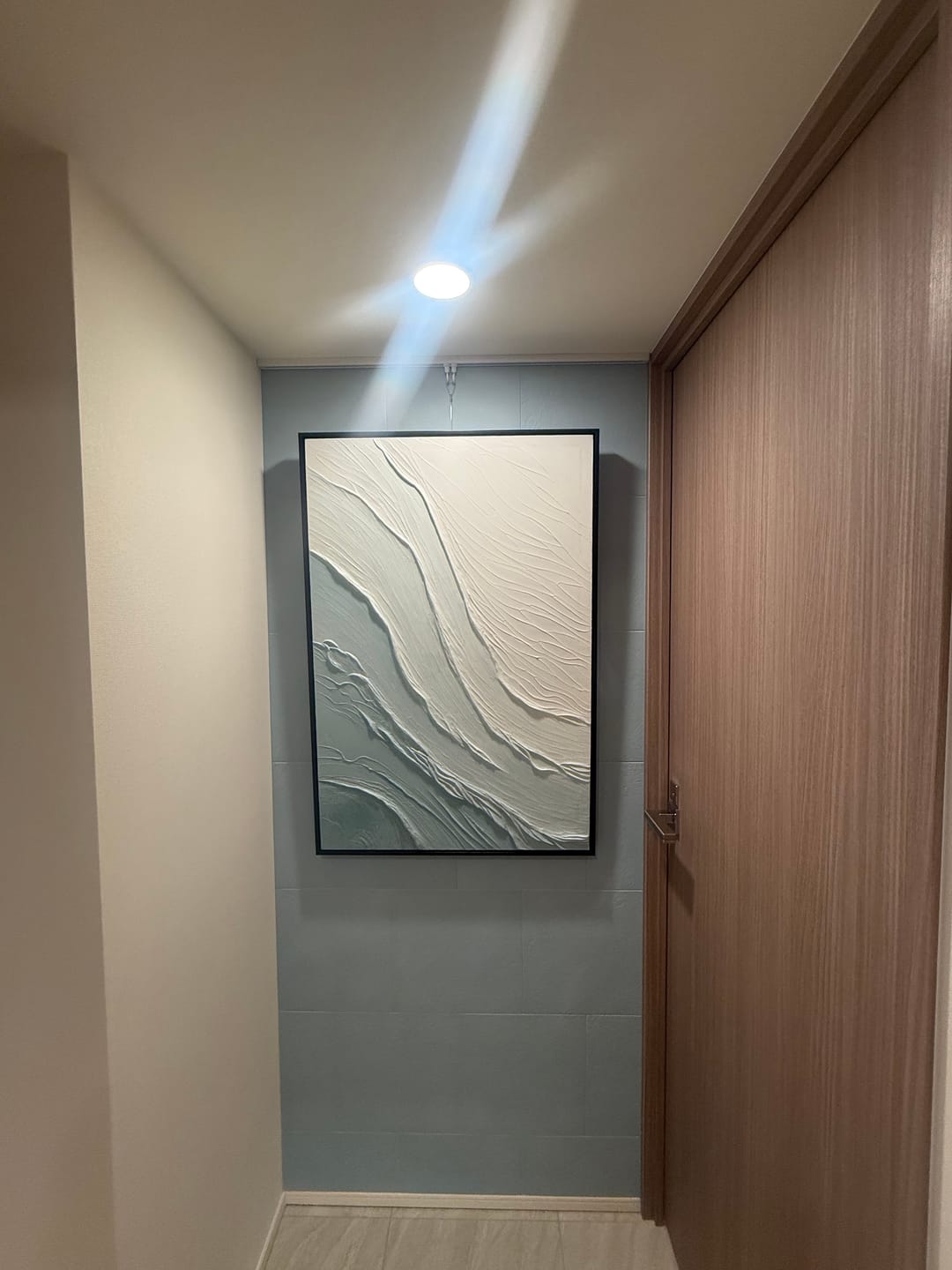 A large framed wabi sabi abstract wall art print with flowing white and gray textured lines, displayed on a light blue-gray tiled wall in a hallway.