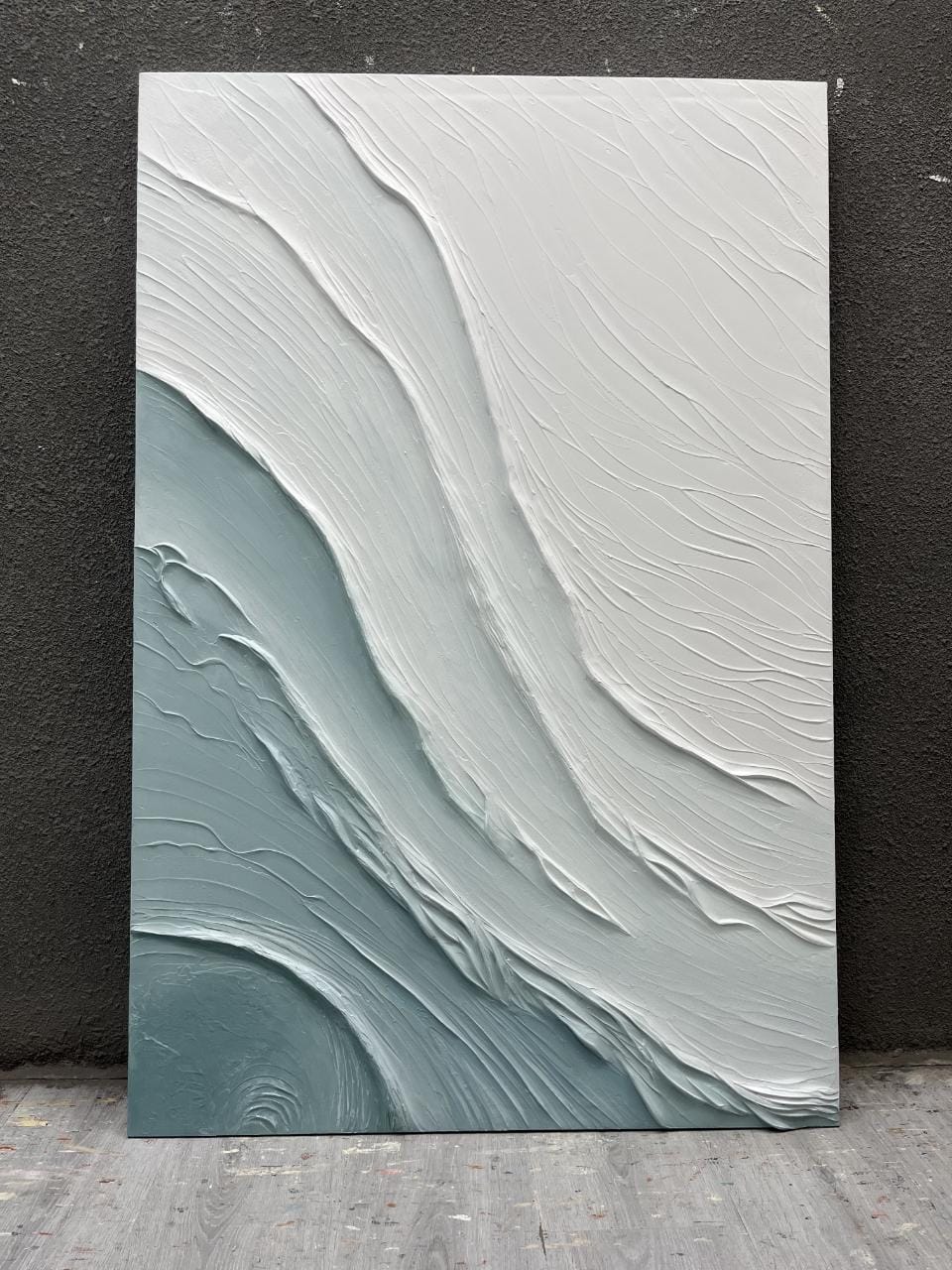 An abstract vertical wall art painting with thick textured brushstrokes in white and pale blue-gray, leaning against a dark wall on a wood floor.