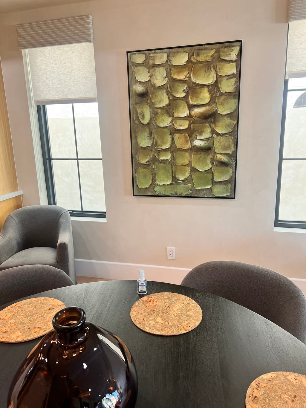 A framed wall art painting with a textured green and gold leaf pattern hangs on a beige wall in a dining room setting above a dining table.