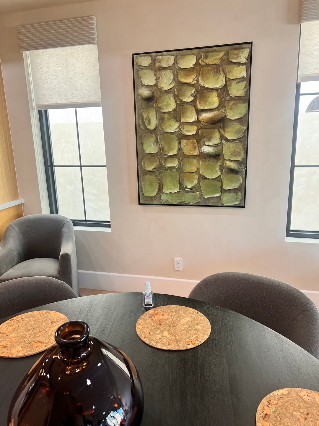 A framed wall art painting with a textured green and gold leaf pattern hangs on a beige wall in a dining room setting above a dining table.