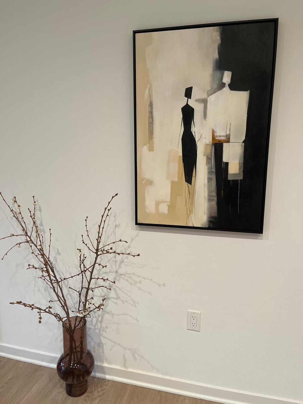 A framed abstract art print featuring black and beige figures, displayed on a white wall above a wooden floor.