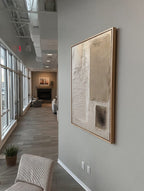 A large abstract framed wall art print with beige, cream, and gray tones hanging on a light gray wall in a modern office hallway.