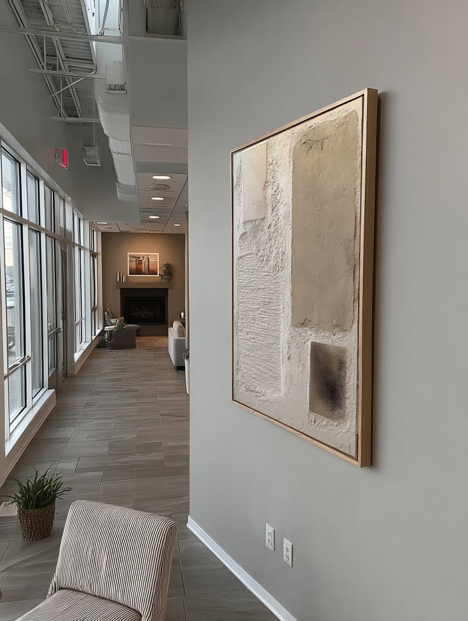 A large abstract framed wall art print with beige, cream, and gray tones hanging on a light gray wall in a modern office hallway.