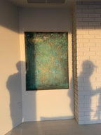 An abstract framed painting with textured teal, green, and gold tones hanging on a beige wall in an outdoor room.
