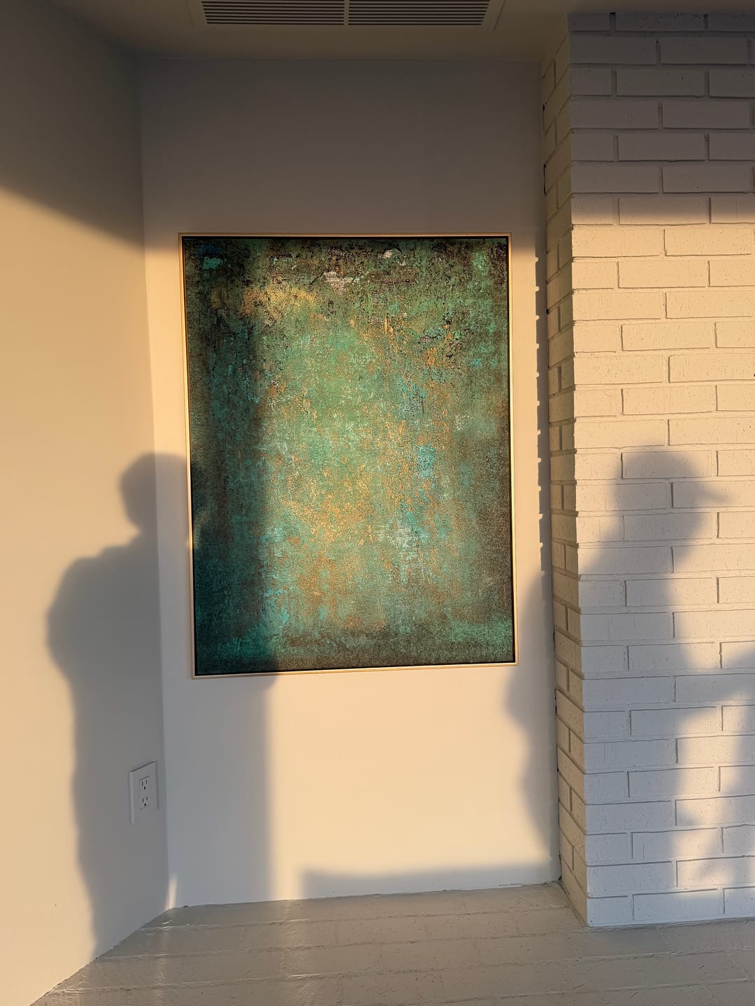 An abstract framed painting with textured teal, green, and gold tones hanging on a beige wall in an outdoor room.