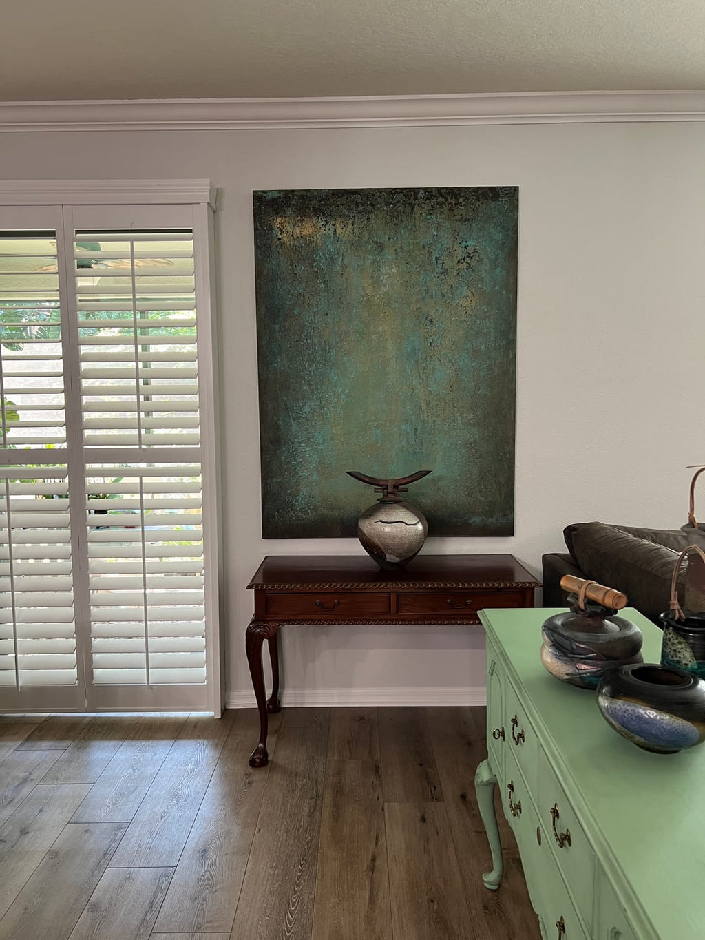 A large vertical textured canvas painting in teal, green, and gold tones, displayed on a white wall above a dark wood console table in a home interior.
