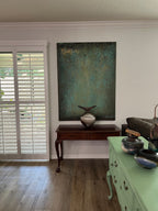 A large vertical textured canvas painting in teal, green, and gold tones, displayed on a white wall above a dark wood console table in a home interior.