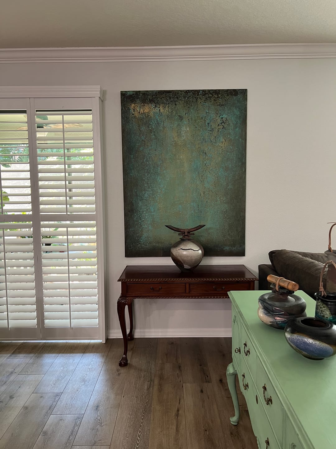 A large vertical textured canvas painting in teal, green, and gold tones, displayed on a white wall above a dark wood console table in a home interior.