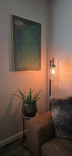 A framed abstract wall art piece with textured teal, green, and gold tones hanging in a living room corner above a small side table.