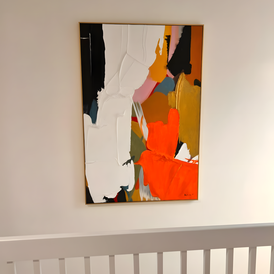An abstract framed painting with bold brushstrokes in white, orange, black, yellow, pink, and blue, hanging on a light beige wall.