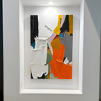 An abstract canvas painting with thick impasto brushstrokes in white, orange, yellow, gray, black, and small accents of pink, blue, and green, displayed in a white recessed gallery wall under a spotlight.